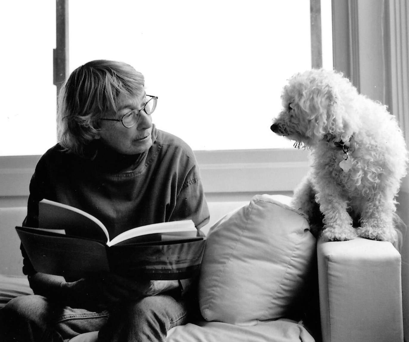 Poet Mary Oliver, wearing glasses reads a book while looking at a small curly-haired dog sitting on the sofa next to her.