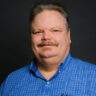 Man with light skin, mustache, and short hair wearing a blue plaid shirt, posing against a dark background.