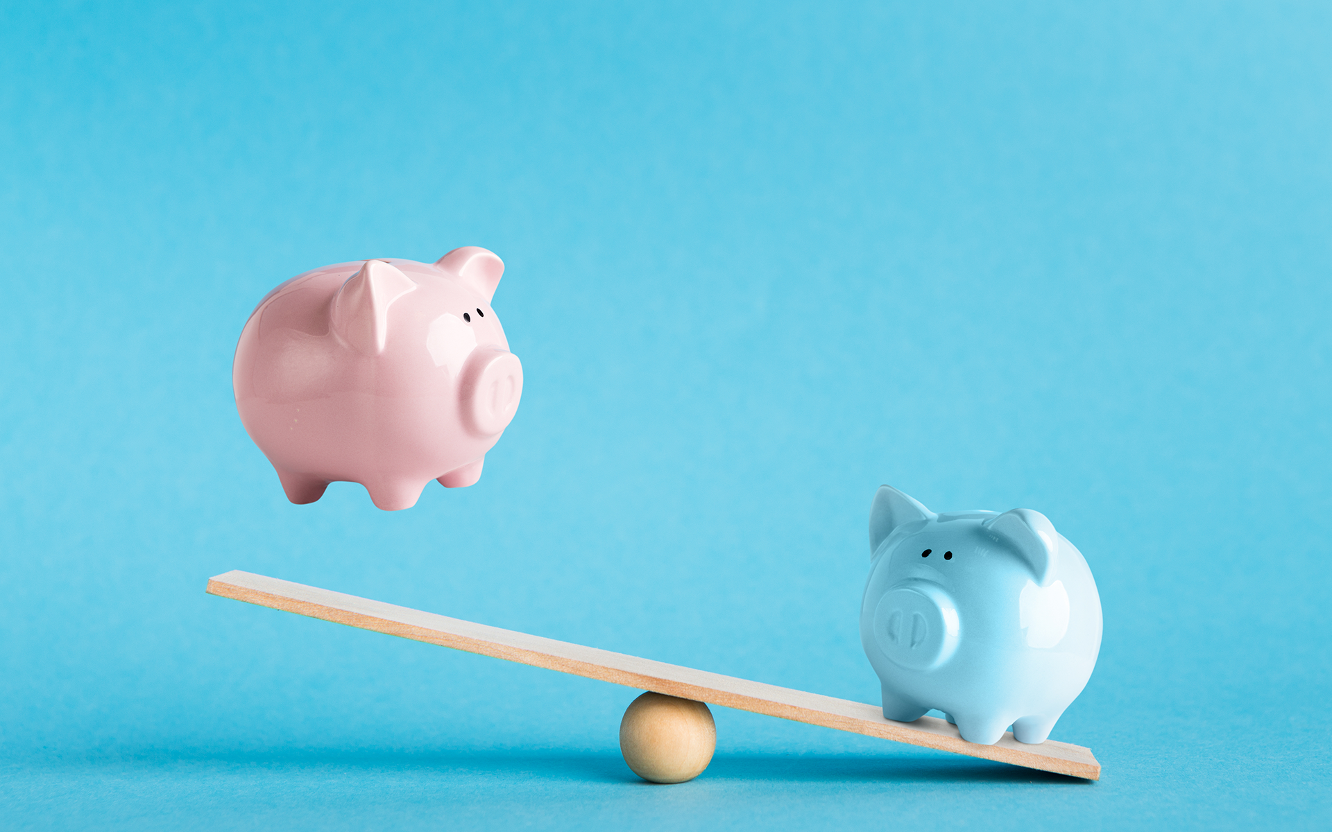 4 Ways to Bring Mindfulness to Your Money Habits — Photo of one blue piggy bank and one pink piggy bank on either sides of a teeter totter with a blue studio background behind.