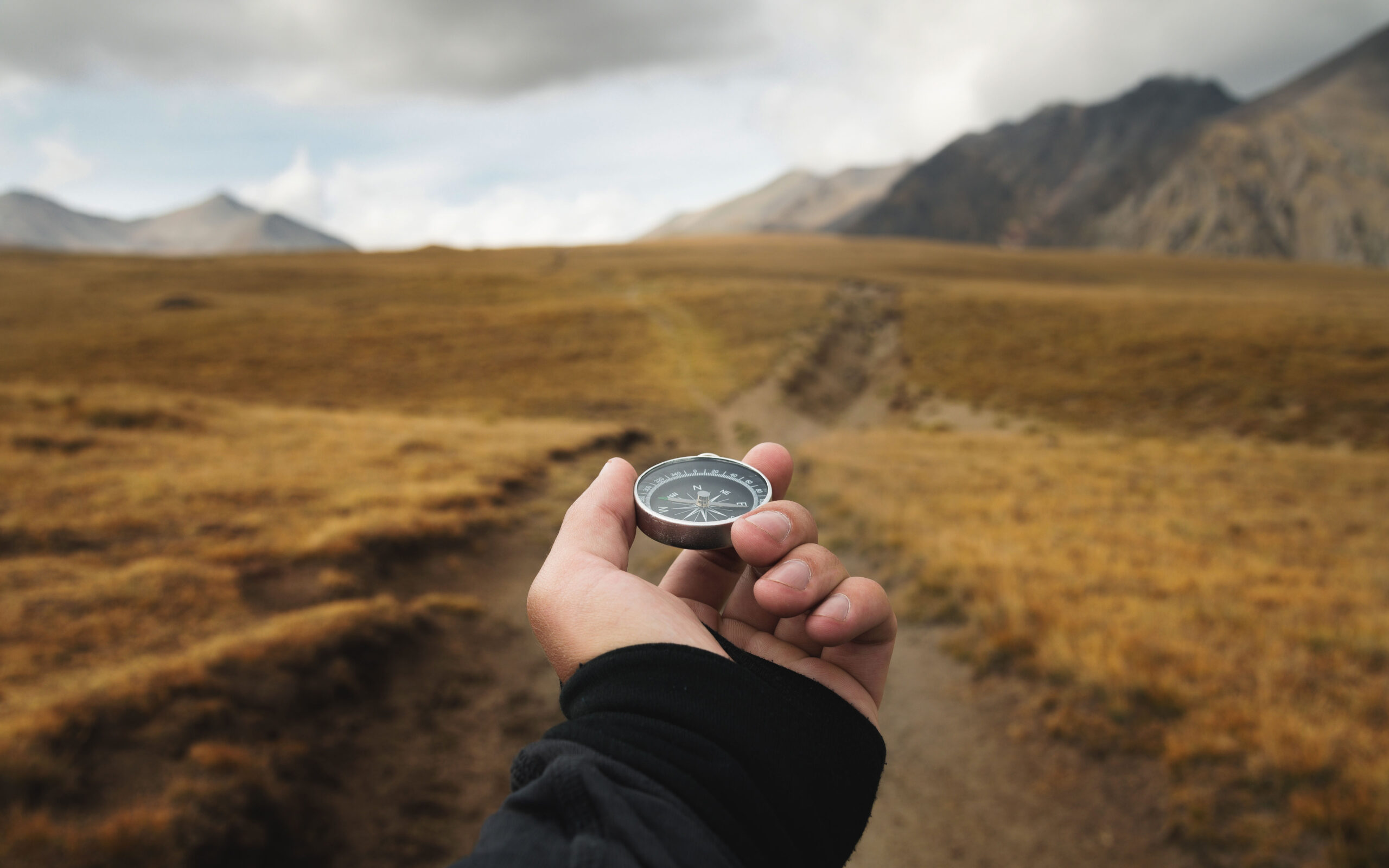 4 Ways to Follow Your Inner Compass - Photo of a person's hand holding a magnetic compass first-person view against the background of a path and mountains