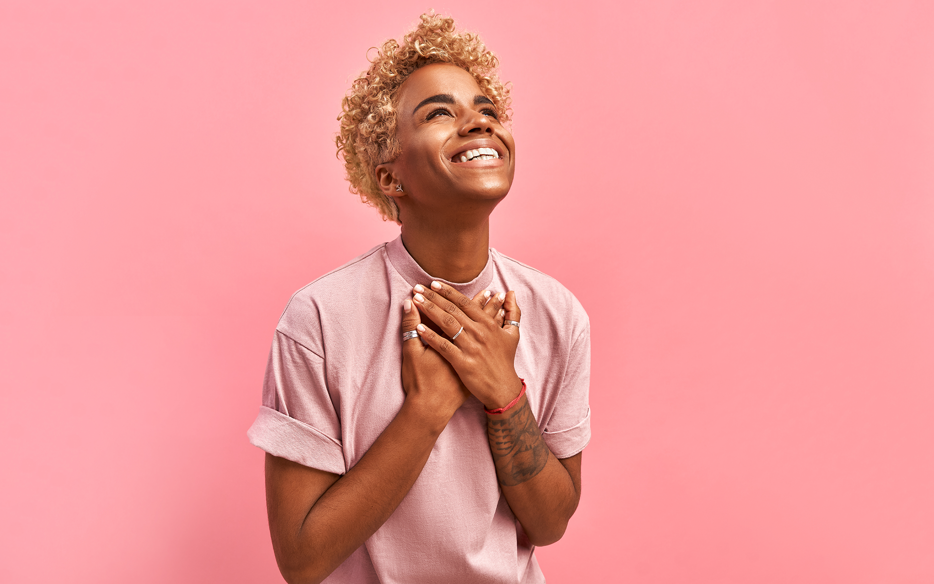 5 Gratitude Meditations to Fill Your Heart with the Joy of Giving Thanks—A smiling Black person with short blonde hair and a pink T-shirt on holds their hands on their chest in front of a pink studio background