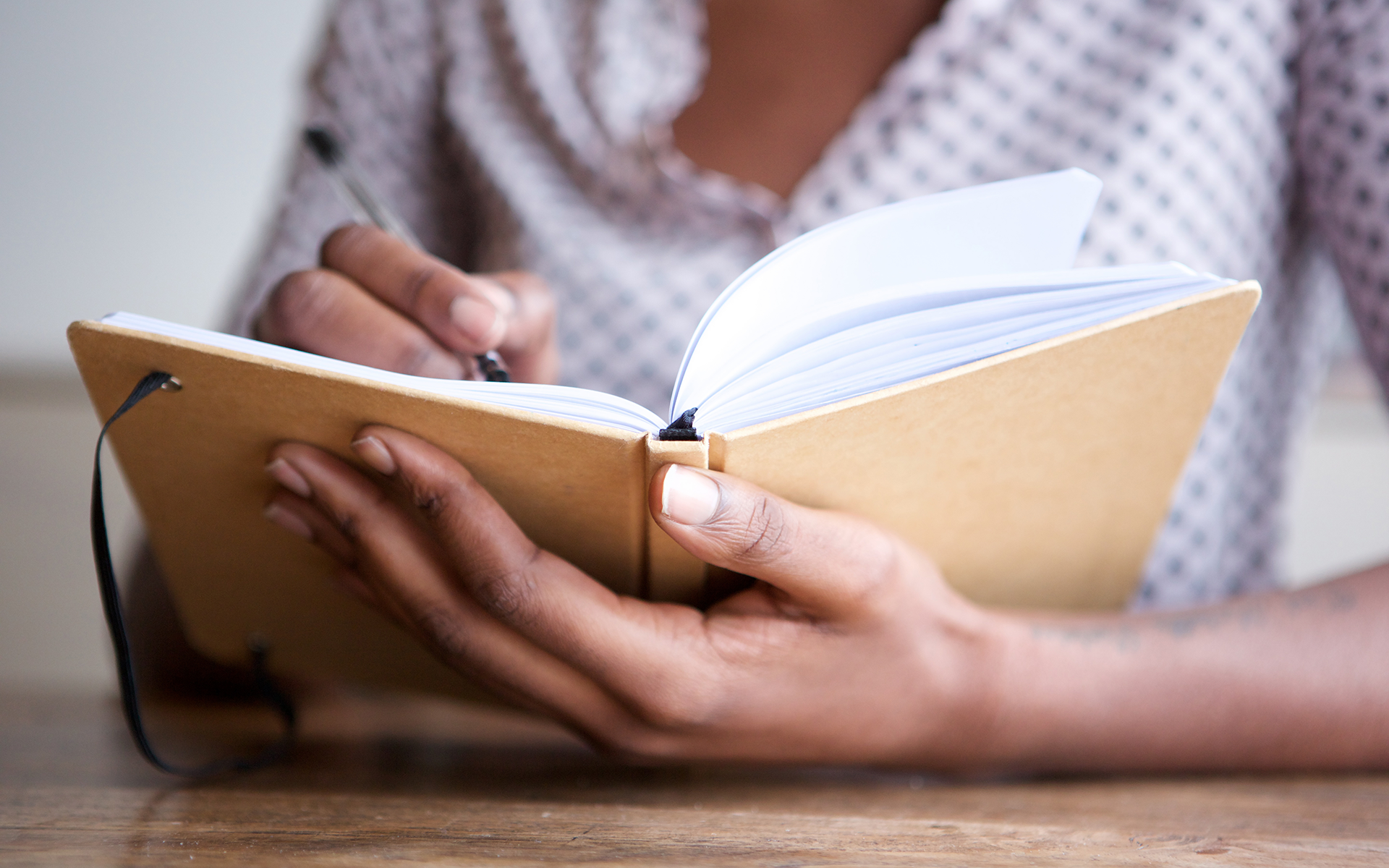7 Mindful Writing Prompts to Foster a Deeper Connection with Yourself—Close-up of a Black woman's hands as she writes in a journal with.a light brown cover.