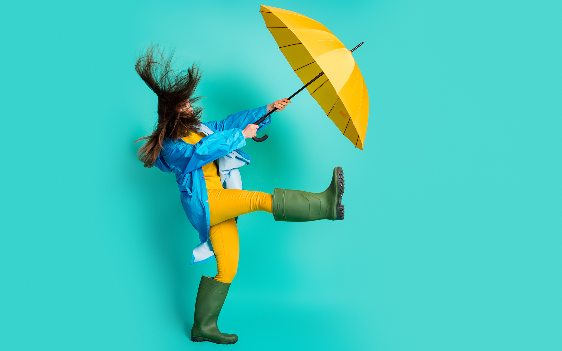 A 12-Minute Meditation to Create Space in a Storm of Emotions—Photo of a woman whose yellow umbrella is caught in the wind with teal studio background.