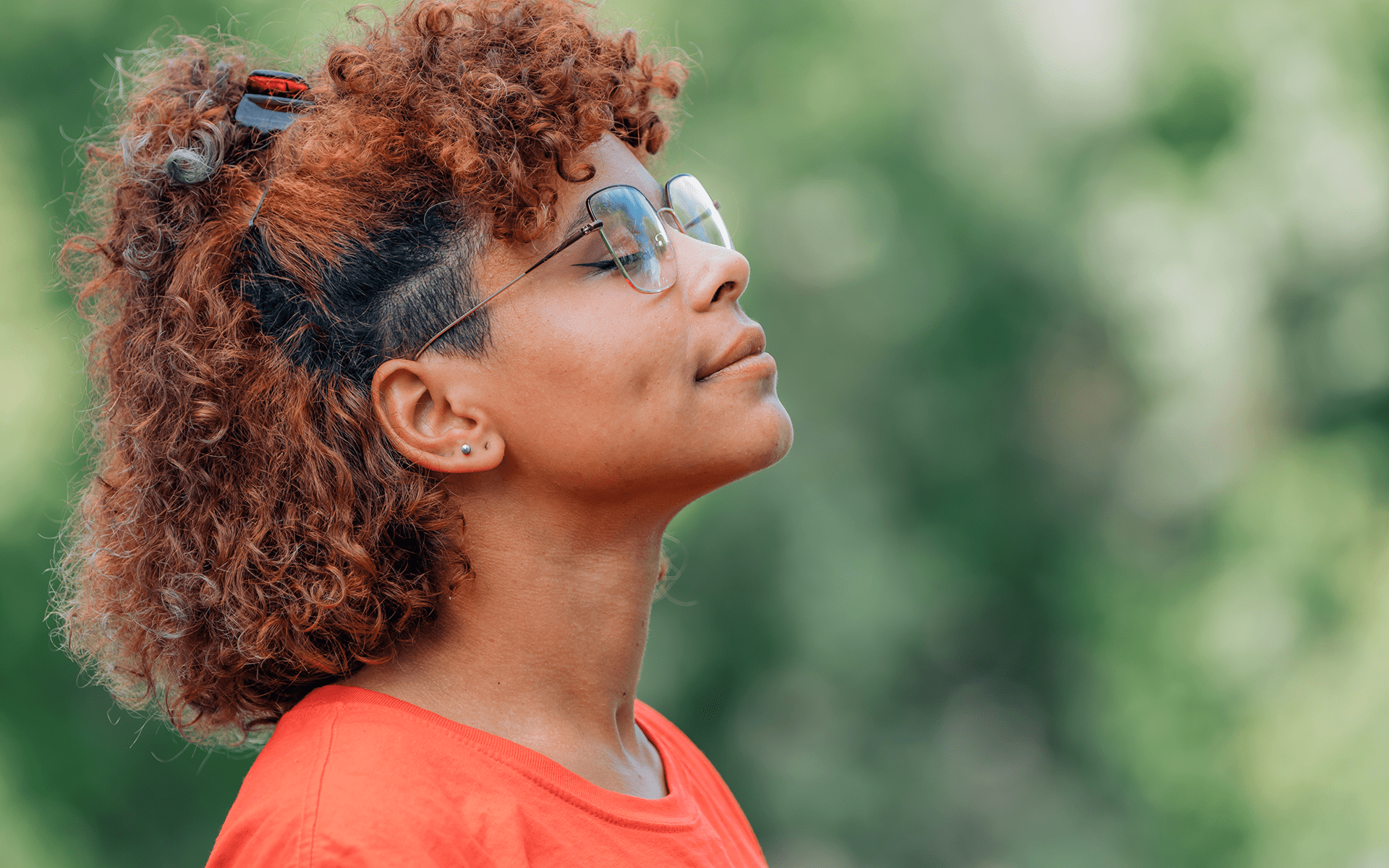 Image of a Black woman with curly brown hair, large glasses, and an orange shirt on stands outdoors with her faced turned upward. She has a peaceful expression on her face.