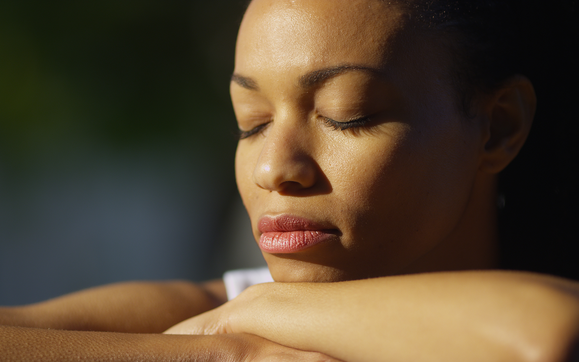 Close-up photo of a Black woman resting her face on her crossed arms with her eyes closed in the sunshine.