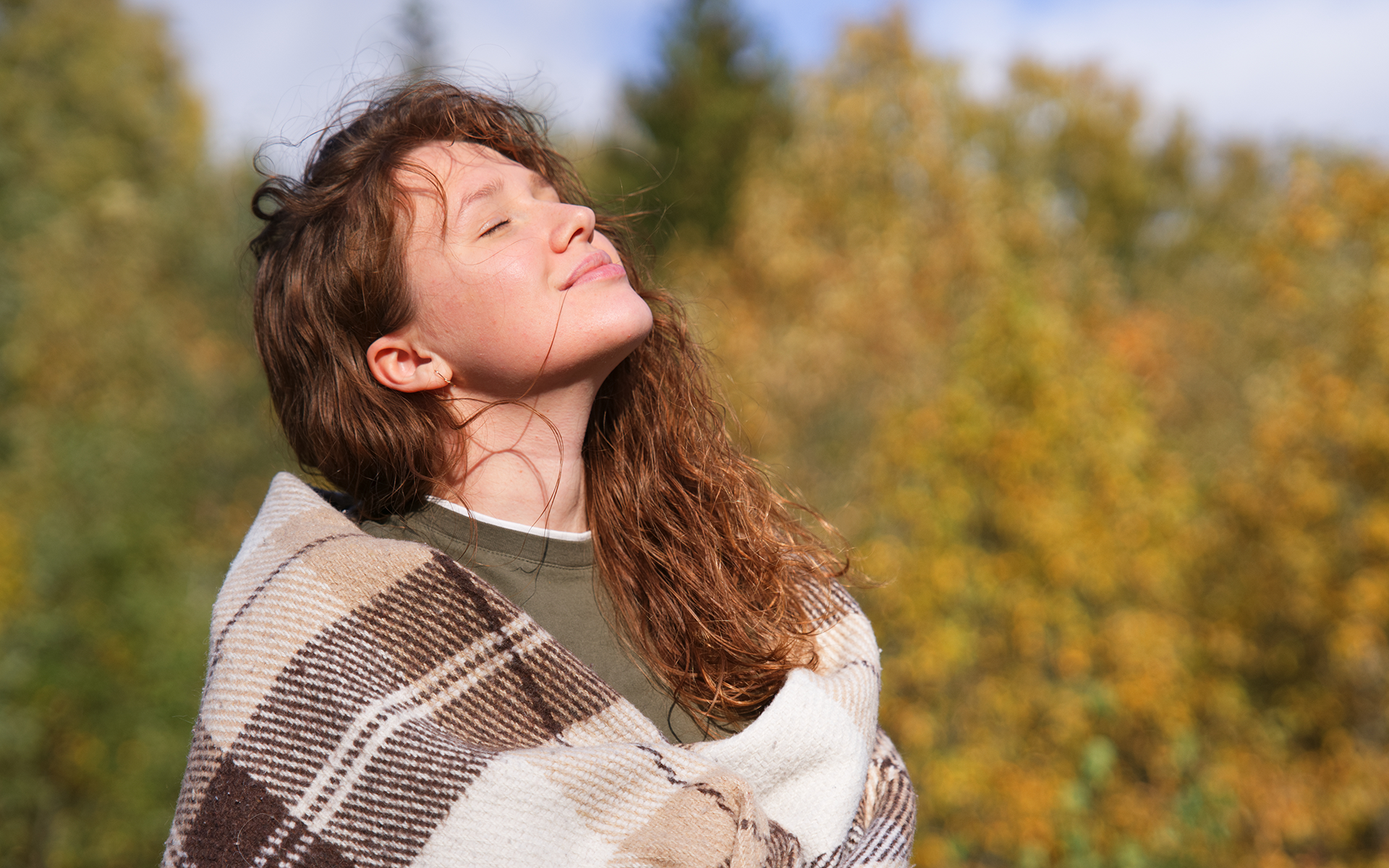 Photo of a white woman with curly brown hair stands outside with her face turned up toward the sun with a peaceful expression, she is wrapped in a brown scarf.