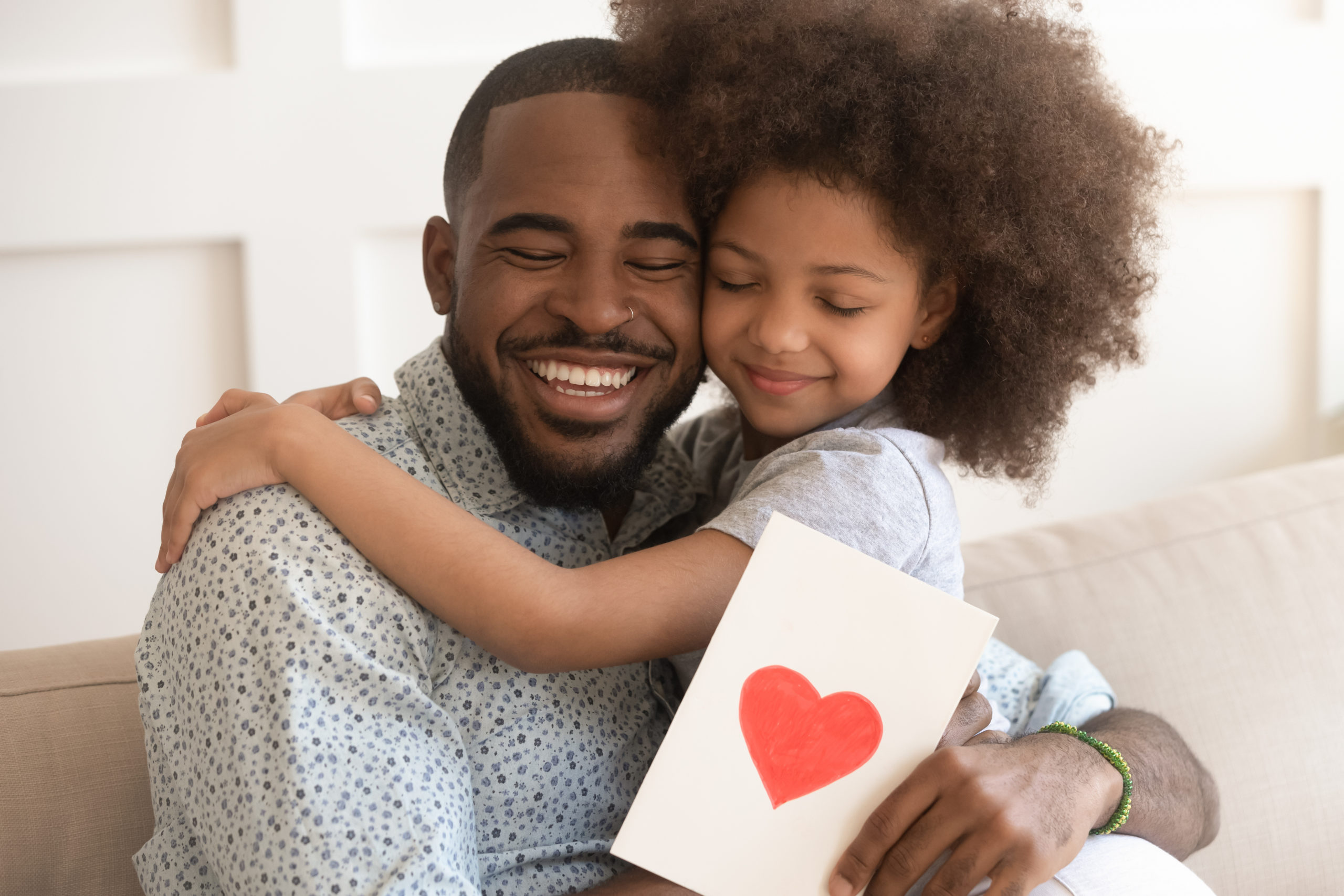 A black man hugging his black son while holding a card with a red heart painted on it