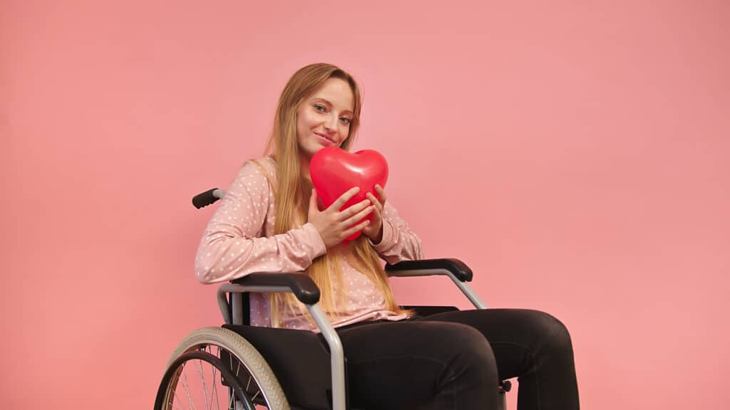 Young white, blonde woman in a wheelchair holds a heart-shaped red balloon with a pink studio background behind her.