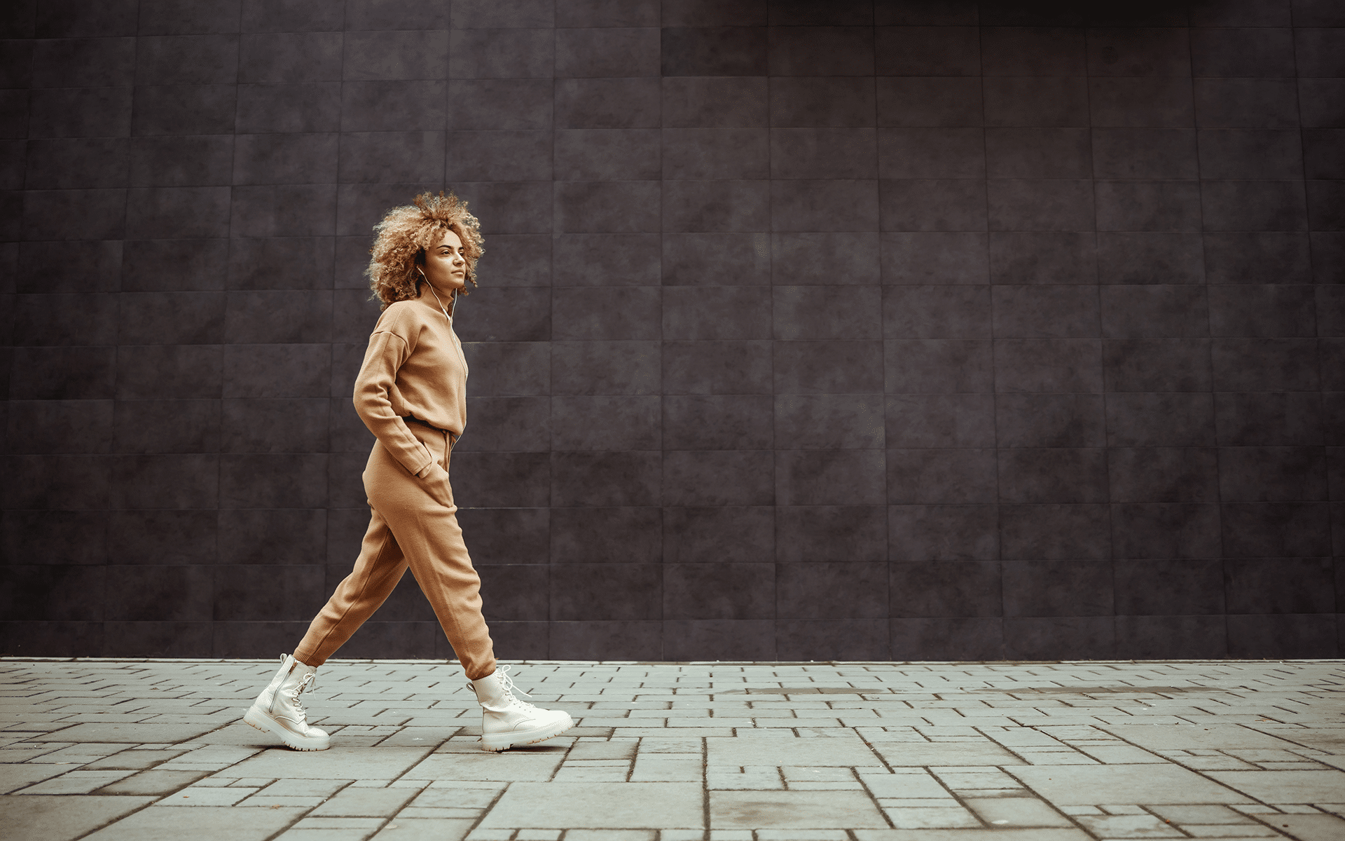 Photo of a woman in a beige tracksuit walking with hands in her pockets and listening music. right way of walking meditation