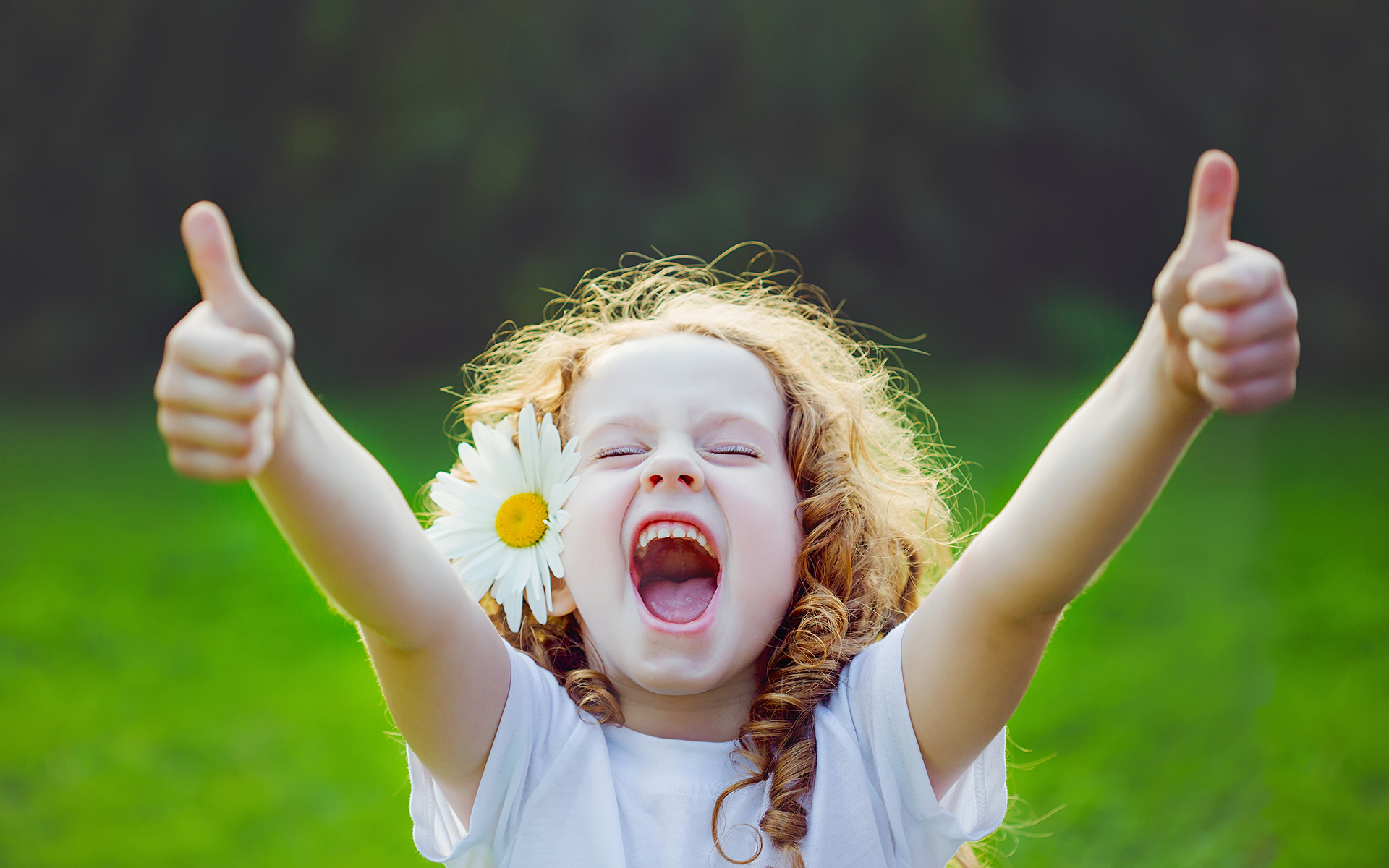 Photo of a little girl with light skin and red curly hair standing outside with a daisy tucked behind her ear with a big smile and giving two thumbs up to the camera.