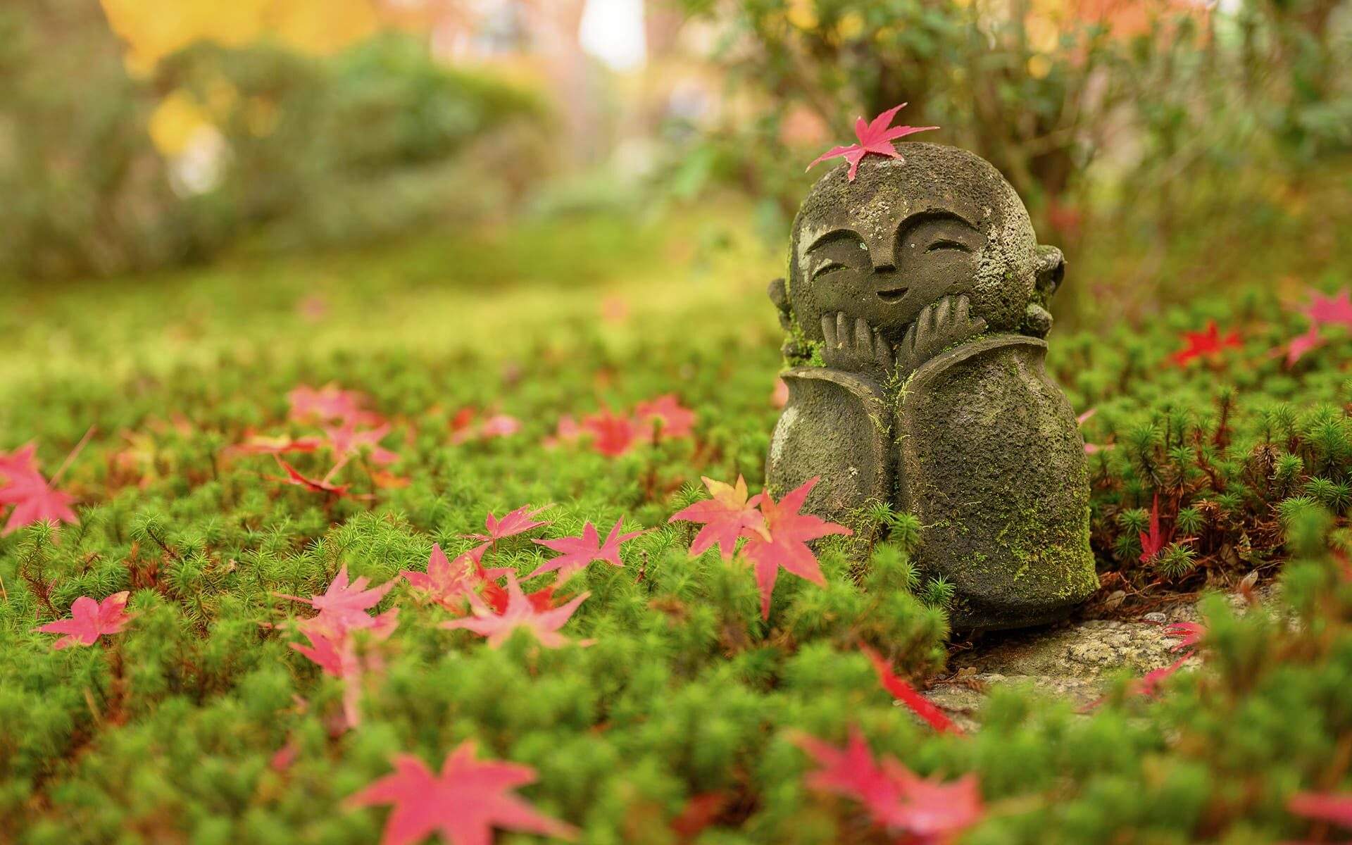 A small stone garden statue of a smiling figure sits in moss, surrounded by red maple leaves for quiet meditation.