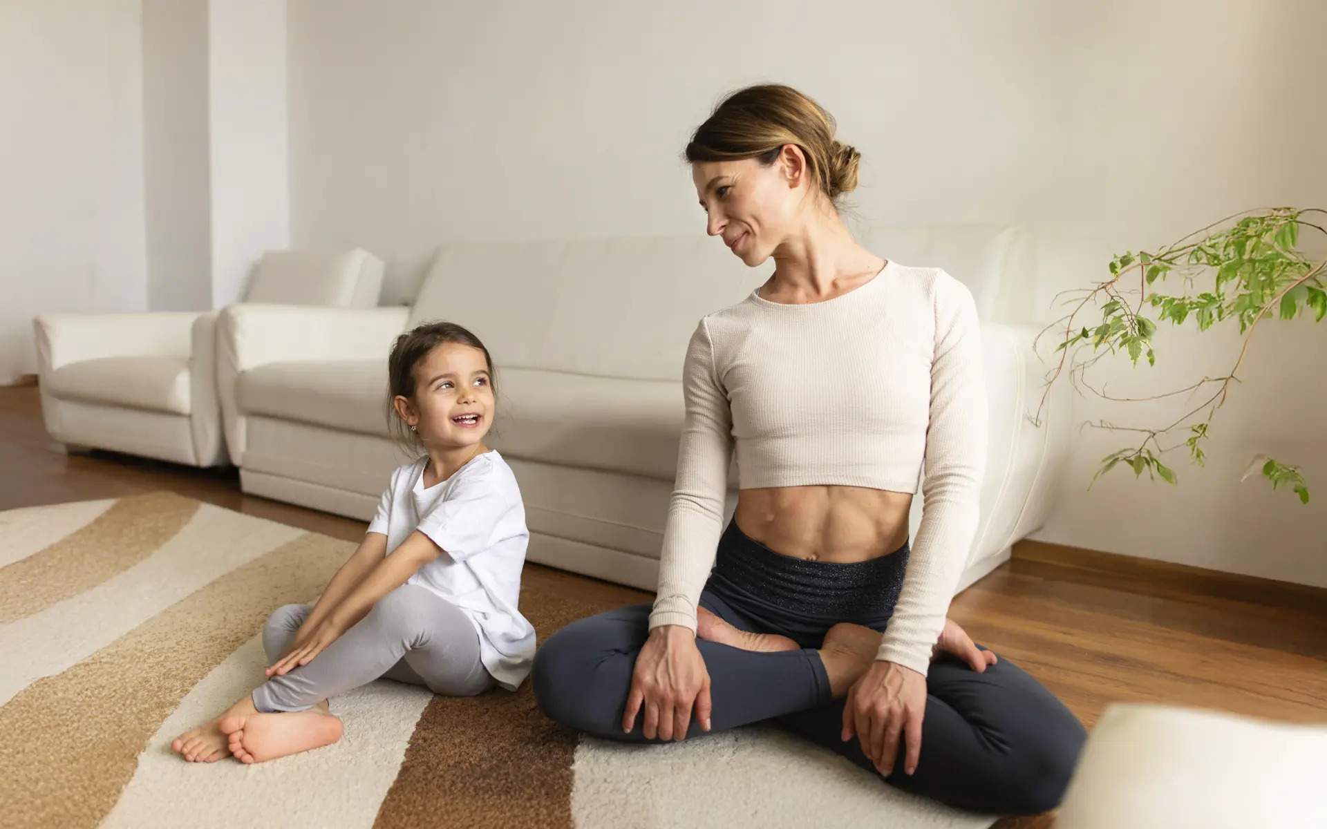 A woman and a young girl sit on the floor in a living room practicing yoga or mindfulness together. The woman is in a cross-legged seated pose with her hands on her knees, while the girl mimics a twist pose, both smiling warmly at each other. They are on a rug in front of a white couch, in a cozy, minimalistic space.