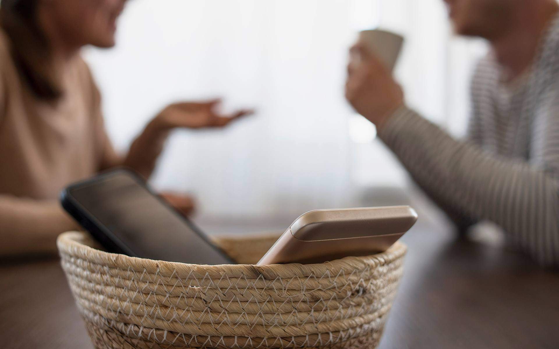 Two smartphones sit in a woven basket on a table as people talk in the background, signaling a screen-free day.
