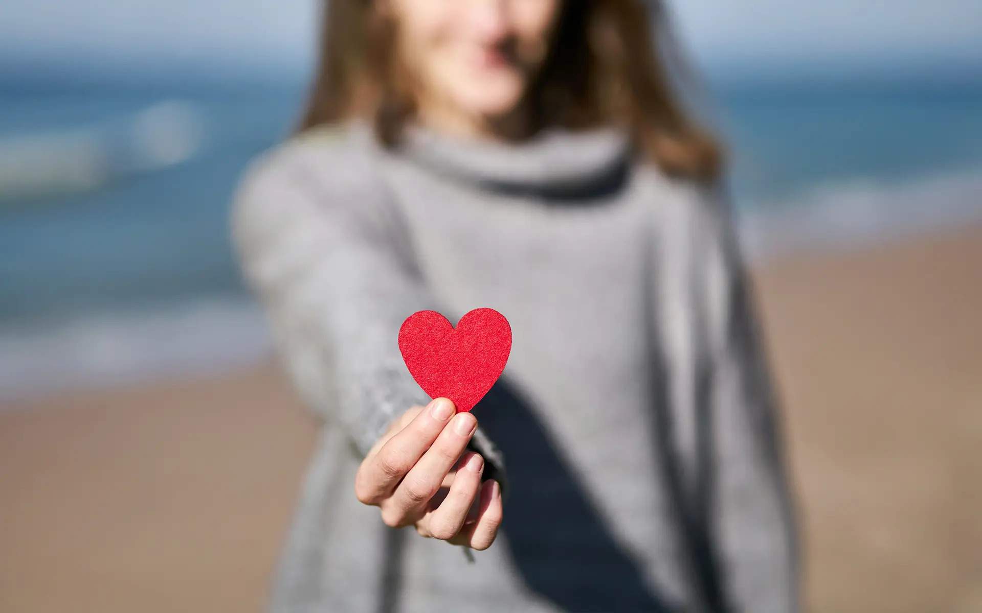 A woman in a gray sweater stands on a beach, holding out a small red heart cutout toward the camera. The background is blurred, emphasizing the heart and her hand in focus.