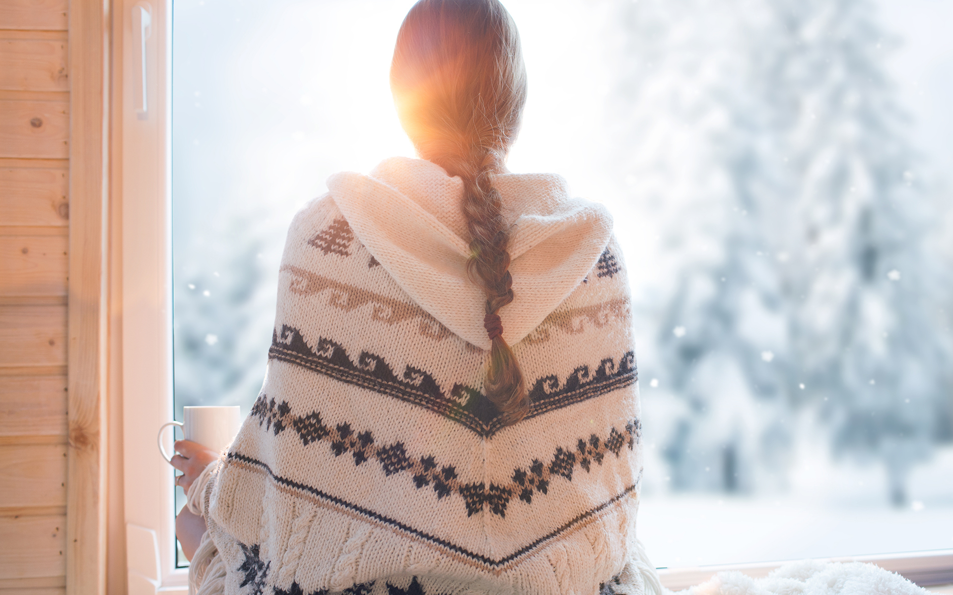 A Guided Practice For Managing Holiday Expectations - Woman wearing nordic style poncho sitting facing a window with cup of coffee, winter woods landscape outside