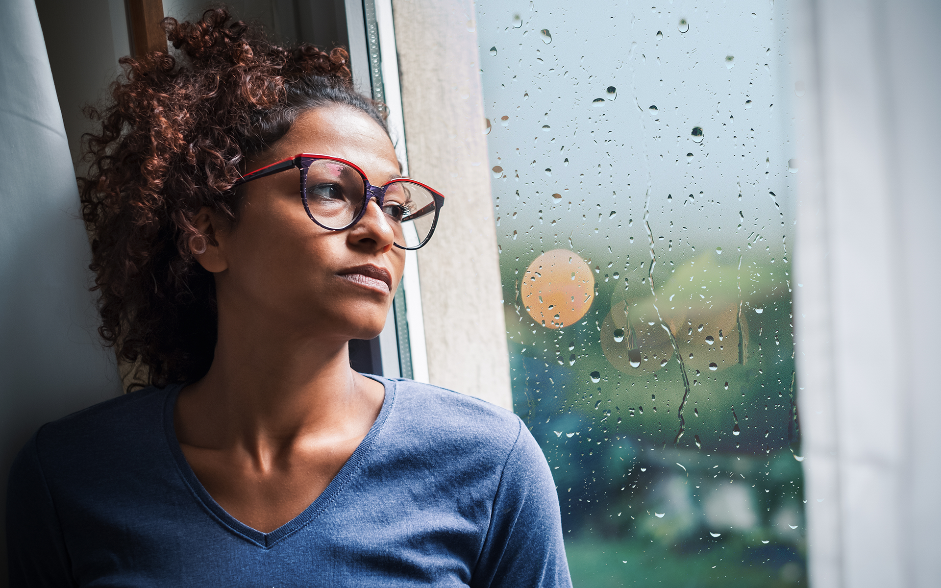 Does Mindfulness Decrease Feelings of Guilt?—Photo of a Black woman wearing glasses and a blue shirt looking out a window at a rainy day.
