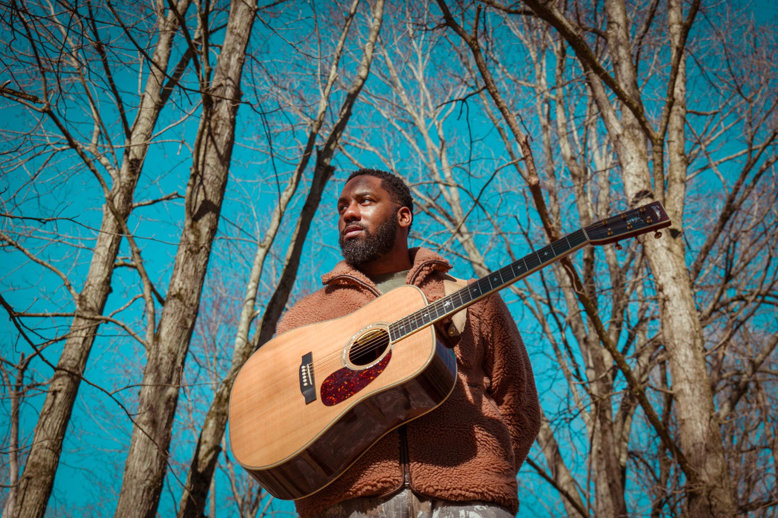 Thaddaeus Washington, mucician and founder of MIND Music, a bearded man stands outdoors before bare trees, holding an acoustic guitar as a tool for his mindfulness practice.