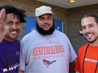 Three men standing outside a brick building, smiling. The man on the left is waving and wearing a purple shirt, the man in the middle is wearing a light gray Baltimore Orioles shirt and white cap, and the man on the right has a beard and is wearing an orange shirt.