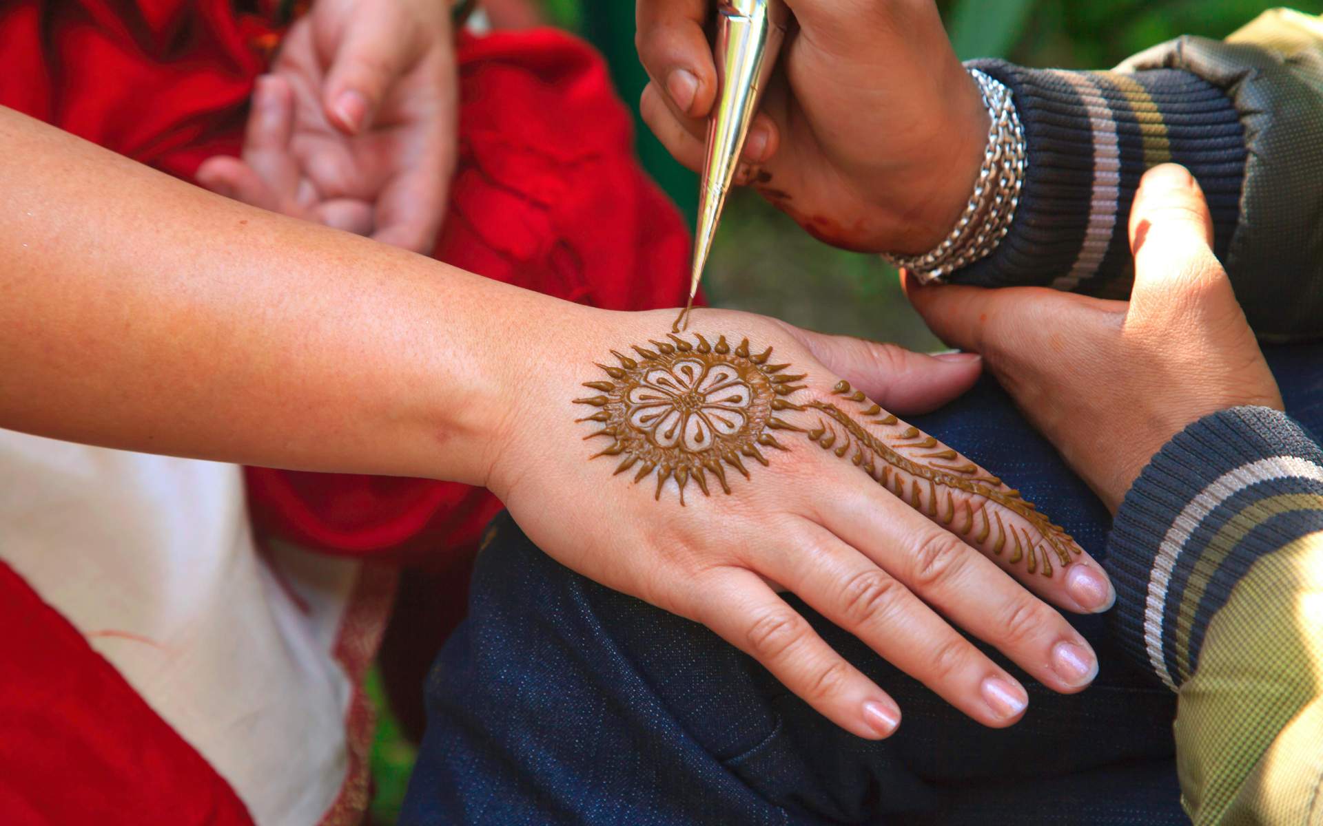 A person is applying a henna design on the back of someone’s hand using a cone. The design features a circular floral pattern with radiating lines and a leafy trail extending toward the wrist. The hands are close together.