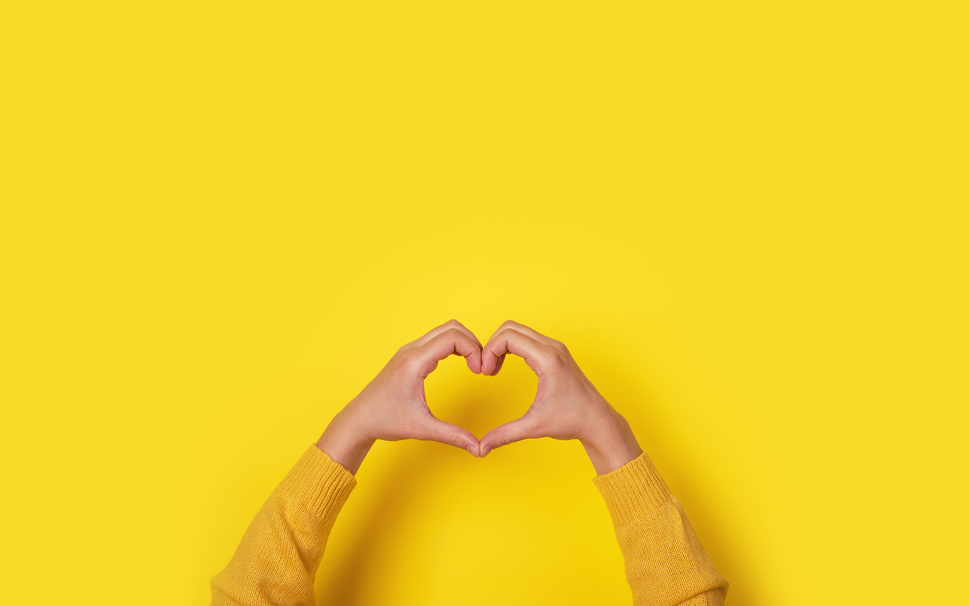How Compassion and Curiosity Help Me to Be Present—Two hands reach up from the bottom of the frame to form a heart with the hands. The person is wearing a yellow long sleeve shirt and the background is yellow.