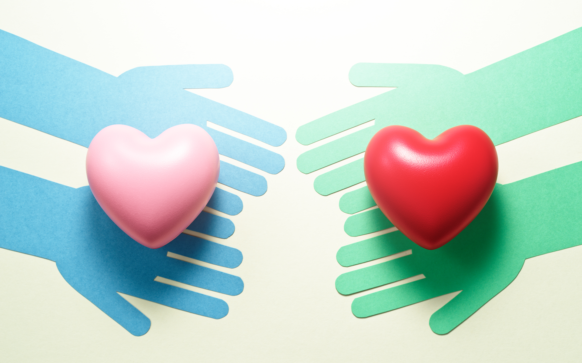 Paper cut outs of two blue hands and two green hands each reaching toward each other with a heart on each set of hands.