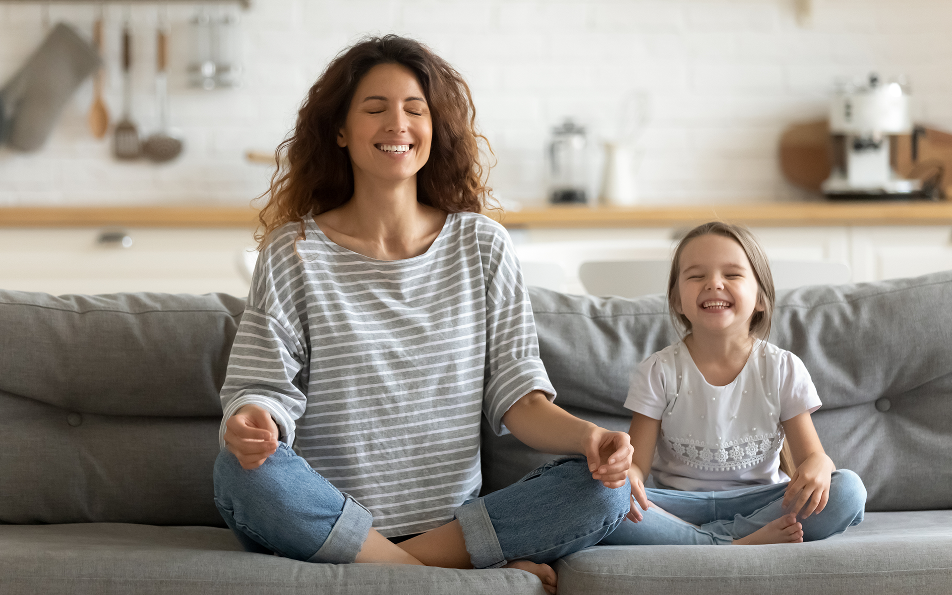 How to Sit in Meditation for Beginners—A woman and a child sit on a couch with their legs crossed and eyes closed. They're both smiling.