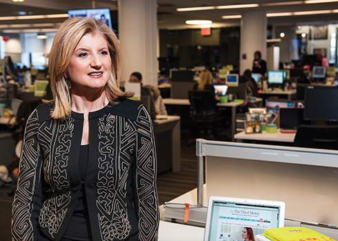 A woman with blonde hair, looking slightly burned out, is wearing a black, intricately-patterned jacket in an office environment. Several desks with computers and office supplies are seen in the background under the bright overhead lighting.