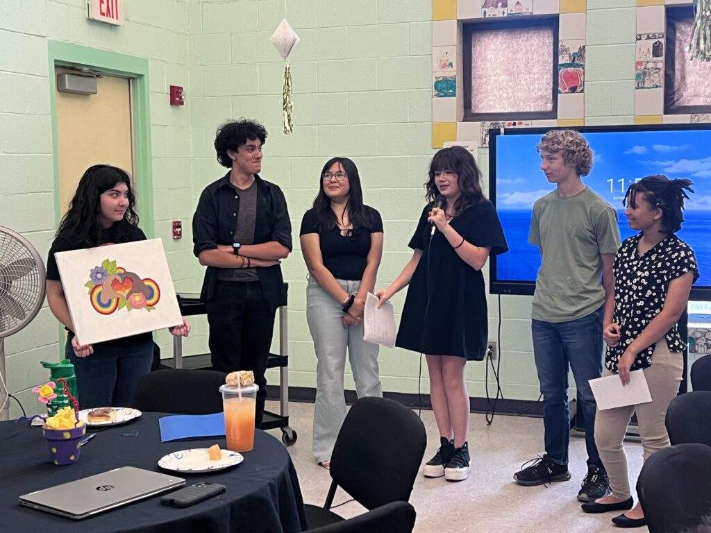 Six young people stand in a classroom—one with artwork, another speaking into a microphone—showing what happens when you Let Them Lead.