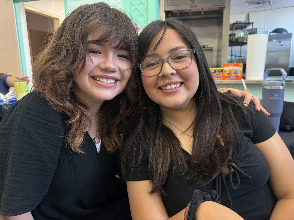 Two young women with long dark hair smiling, sitting close together indoors, arm around each other—Let Them Lead with friendship.