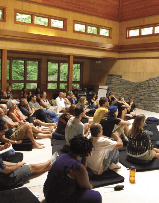 A retreat guide leads a group sitting cross-legged on cushions in a sunlit room with large windows and wooden walls.