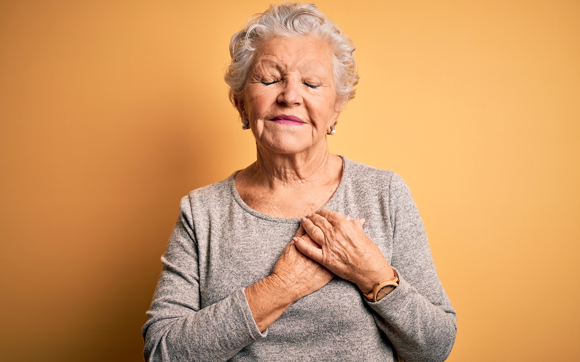 Senior beautiful woman wearing casual t-shirt standing over isolated yellow background smiling with hands on chest with closed eyes and grateful gesture on face.