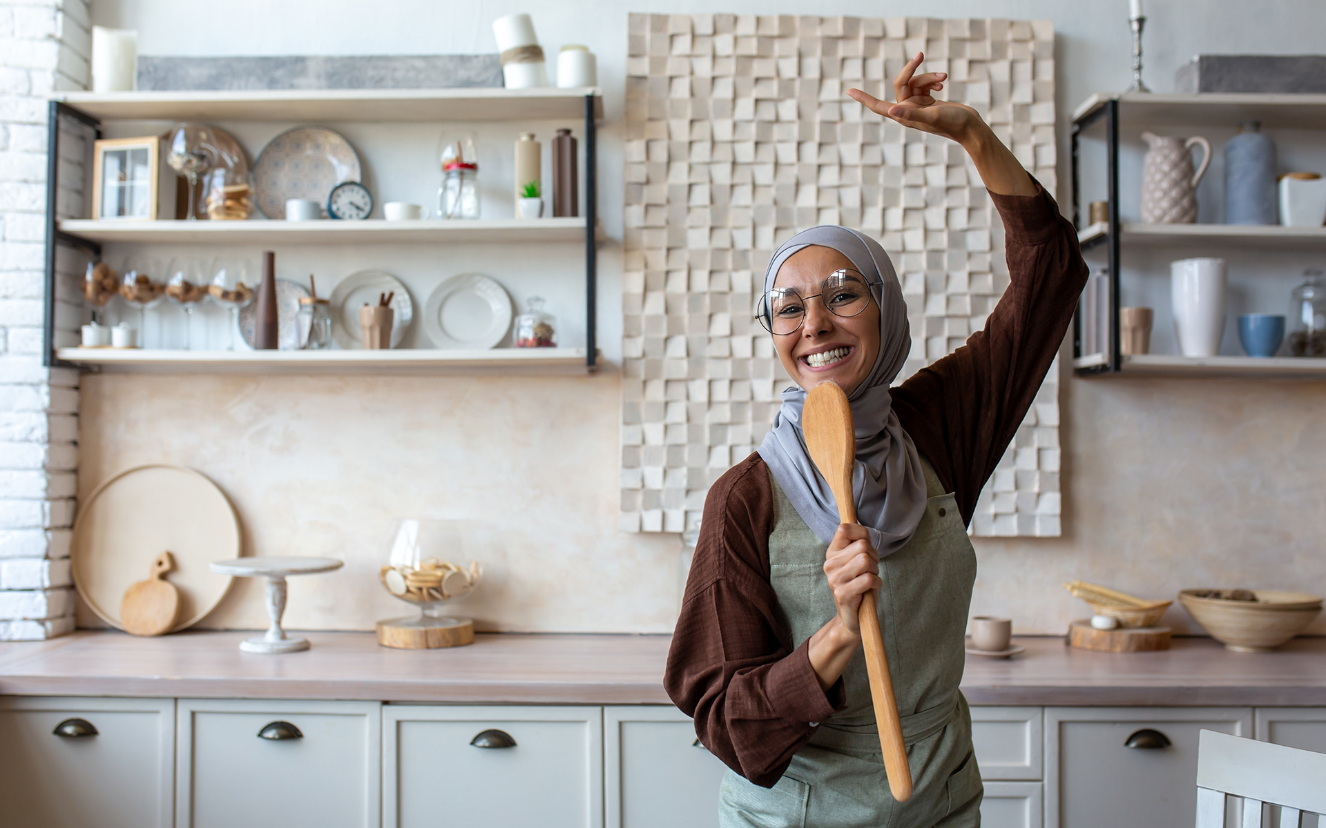 Photo of a woman in a hijab dancing in a kitchen with a joyful expression.