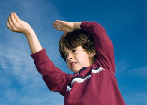 A young child with brown hair wearing a red long-sleeve shirt is standing outdoors against a clear blue sky. The child is raising both arms, one bent and the other extended, and appears to be playfully reaching or pointing at something above.