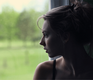 A woman with her hair tied up in a messy bun gazes out a window. The background is blurred, showcasing green foliage and trees. Her expression appears contemplative as soft light illuminates her face.