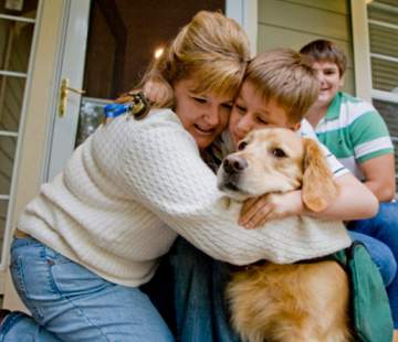 A woman and two children are sitting on a porch, hugging a golden retriever. The woman and younger child, perhaps finding a moment of mindfulness together, have their arms wrapped around the dog, smiling warmly, while the older child sits behind them, also smiling.