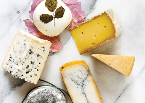 A variety of cheeses arranged on a marble surface. The selection includes blue cheese, a round cheese adorned with a leaf, a wedge of yellow cheese with holes, a slice of hard cheese, and other artisan cheeses, some with rinds and distinctive textures.