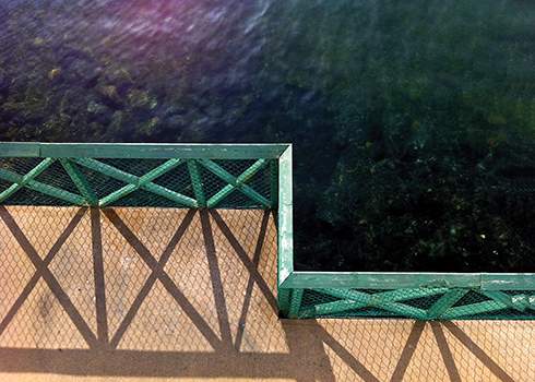 A green metal fence casting crosshatched shadows on a tan concrete surface beside a body of dark, clear water. The fence angles at a right corner, almost signaling a stop, dividing the shot with water on one side and the concrete on the other.