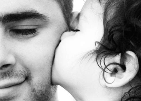 A close-up black and white photograph of a child giving a kiss to an adult's cheek. The adult's left eye is closed, and they are smiling slightly. The child has curly hair, and both individuals' faces show tenderness and affection, capturing a perfect moment for a day for dads.