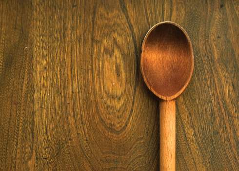 A wooden spoon resting on a wooden surface. The spoon's bowl is oriented to the right, and the handle is toward the left. The wood grain of the surface is distinct and patterned, complementing the texture and color of the spoon.