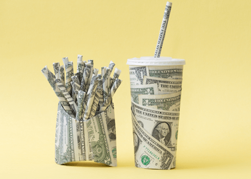 A fast food fry container and drink cup, both covered with US dollar bills, sit against a yellow background. The container is filled with rolled-up dollar bills, mimicking fries, and the cup has a rolled-up dollar bill as a straw.