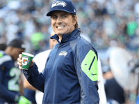 A person wearing a Seattle Seahawks cap and jacket, holding a Gatorade cup, smiles at the camera on a football field. Other team members can be seen in the background, with a blurred crowd in the stands.