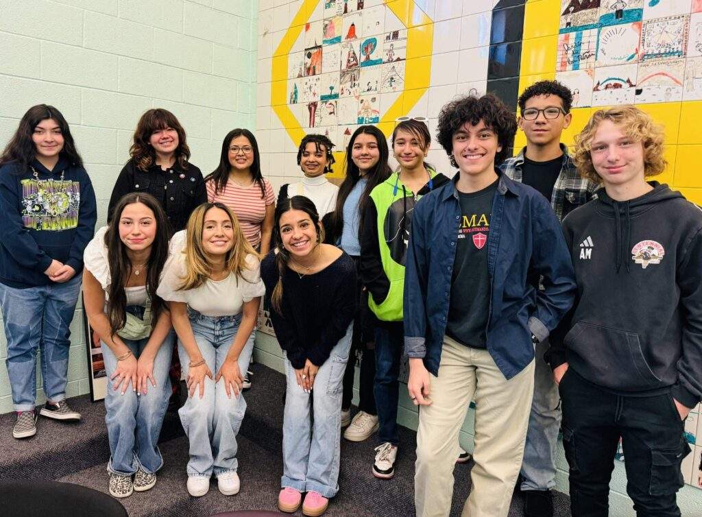 Thirteen smiling teens pose indoors in front of art and a big yellow O, showing the spirit of "Let Them Lead.