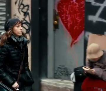 A woman bundled in a black jacket and scarf walks past a wall adorned with graffiti, including a large red heart. In the background, another person, wearing a beige hat and coat, sits on the sidewalk focused on something in their hands.