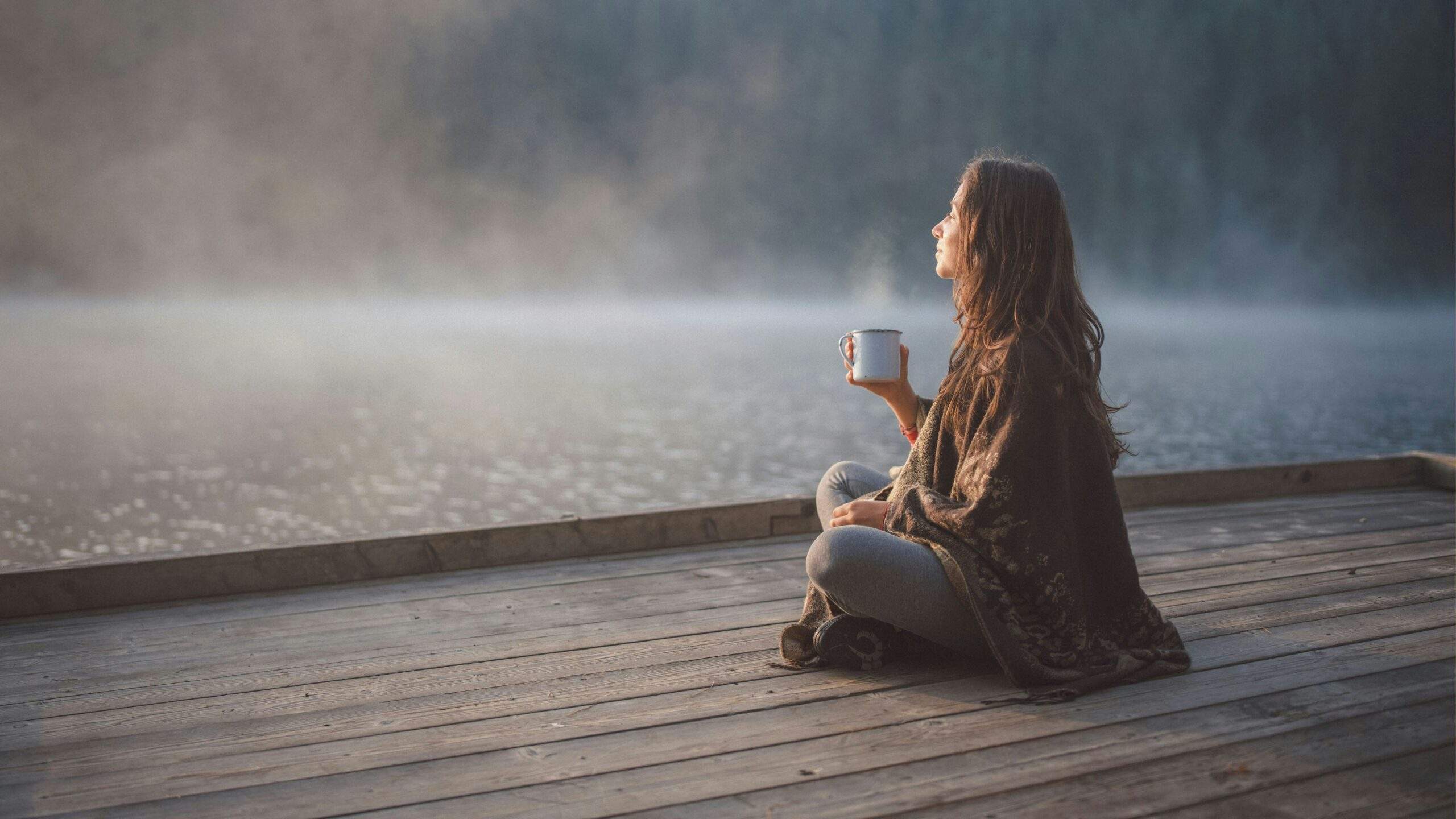 Woman with cup of tea sits on a misty dock at sunrise.