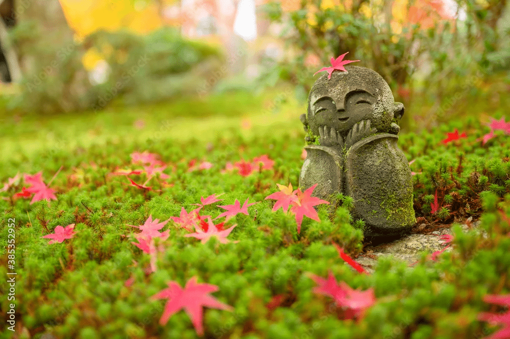 A small stone statue for garden meditation, a smiling figure sitting in mossy ground covered in red autumn leaves.
