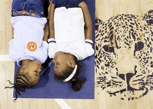 Two children lie on a blue mat on a gym floor, facing each other, with a large painted leopard's head on the floor beside them. They appear to be in relaxed and playful postures.