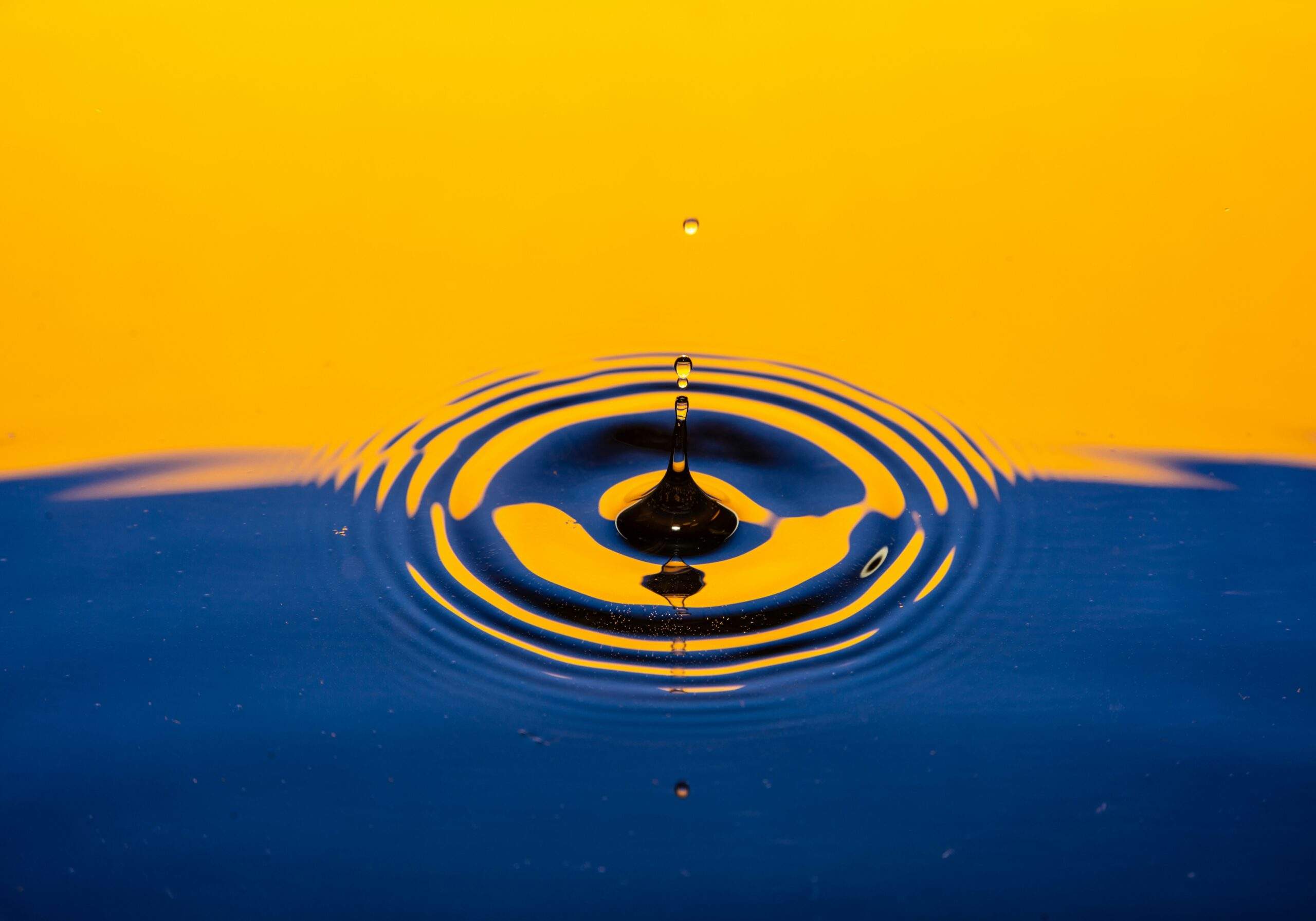 A water droplet splashes, creating a ripple effect—symbolizing personal growth—on a blue surface with a vibrant yellow background.