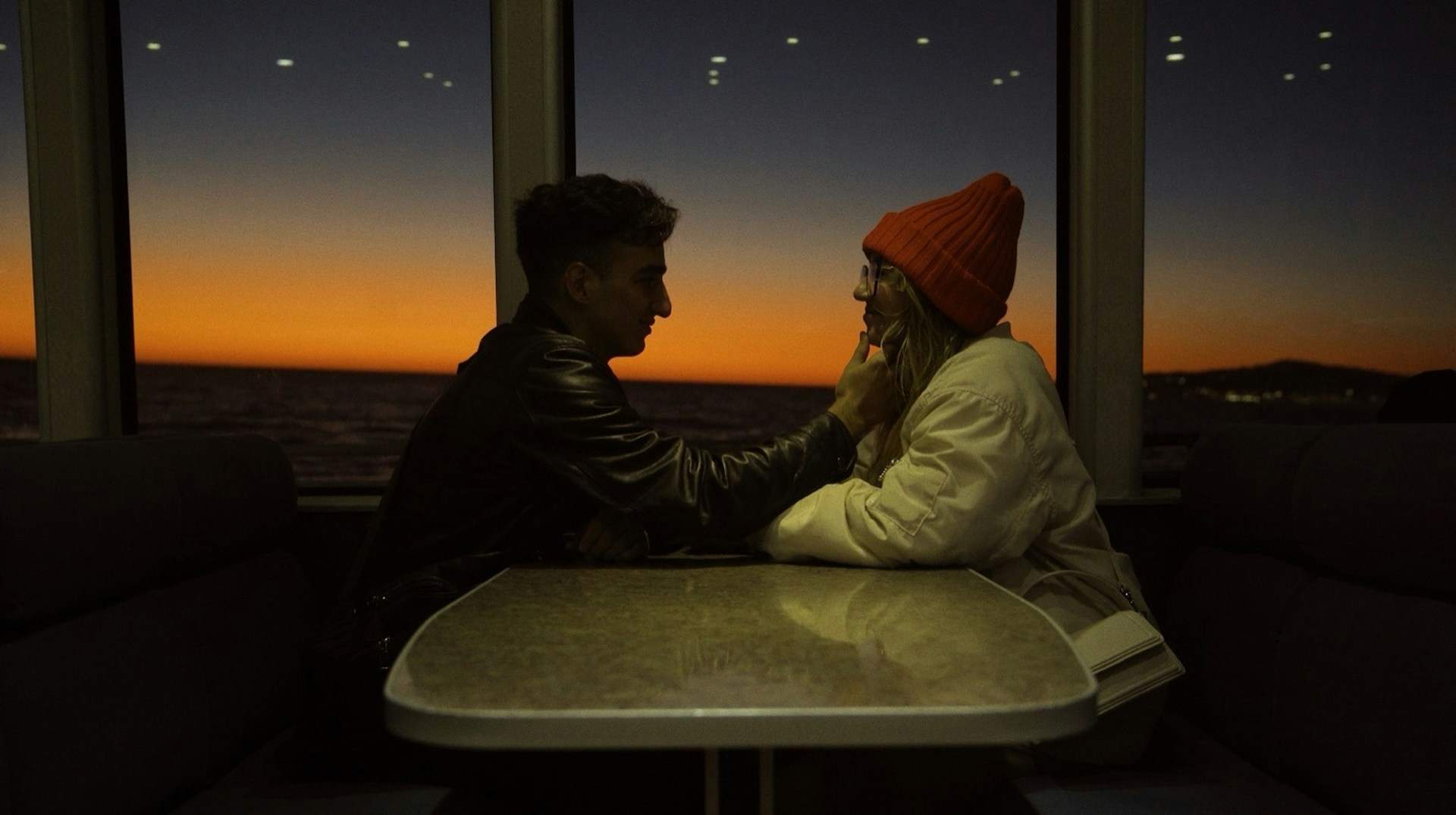 A couple sits across from each other at a table, gazing intently, as if learning how to fall in love beneath a glowing sunset.
