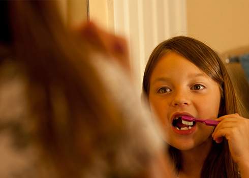 A young girl with long brown hair is brushing her teeth in front of a mirror. She is using a pink toothbrush and appears to be looking at her reflection. The background is softly lit, creating a warm atmosphere.
