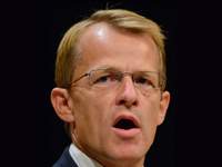 A man with short blond hair and glasses speaks with an open mouth against a dark background. He wears a dark suit jacket and a white shirt.